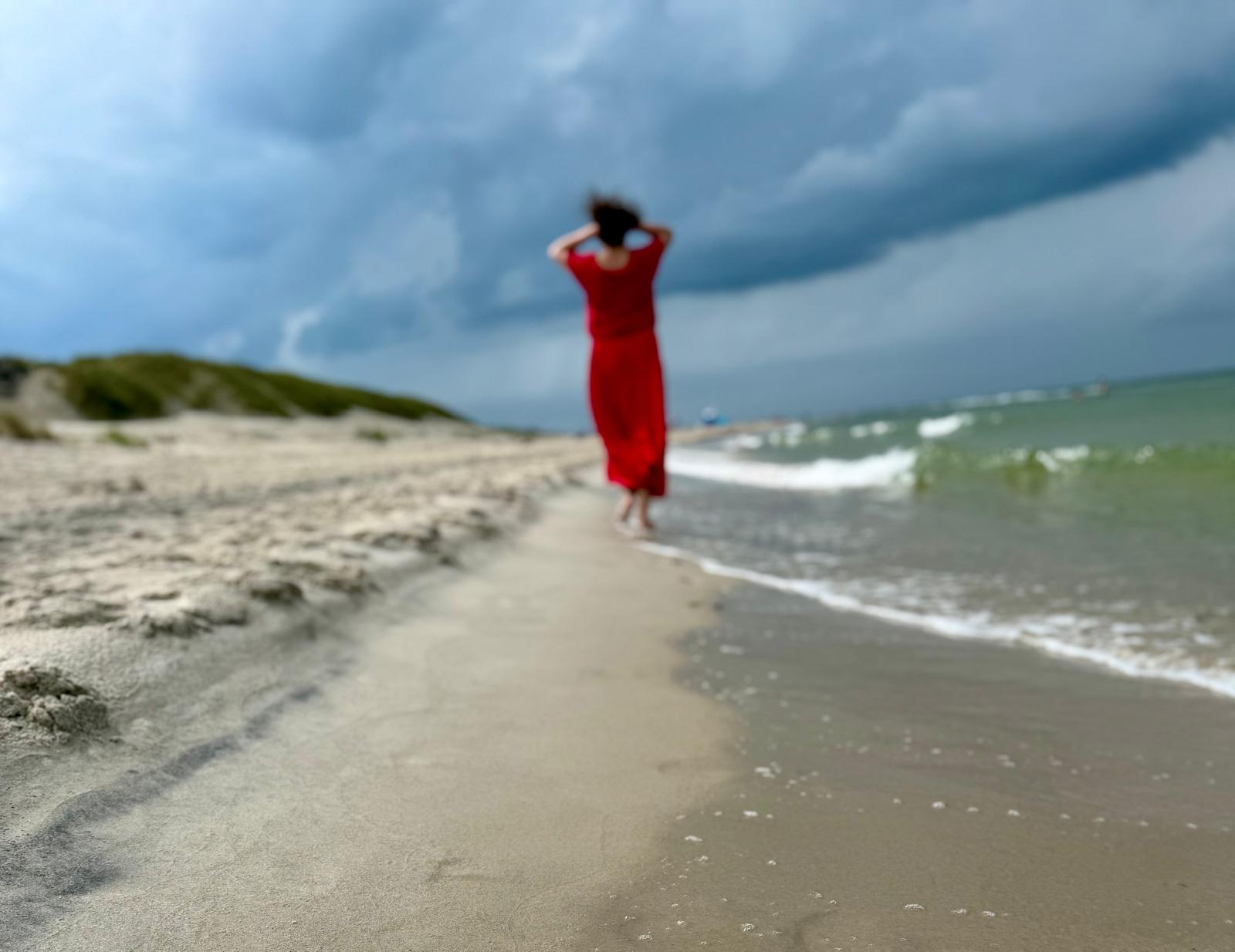 Karolina w czerwonej sukni stojąca na plaży, odwrócona ku morzu